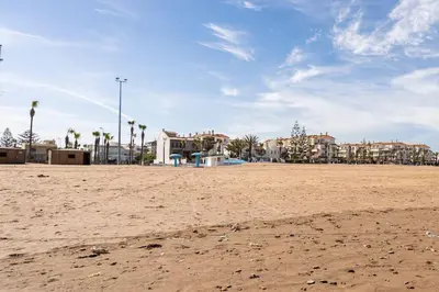 a sandy beach with buildings and palm trees in the background at New appartement next to the beach Sidi Rahal in Sidi Rahal