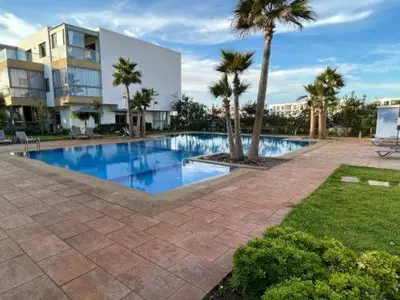 a swimming pool with palm trees in front of a building at Blue beach Sidi Rahal in Le Polo