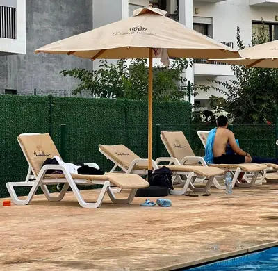 a man sitting in chairs under an umbrella next to a pool at appart luxueuse a rabat hay riad avec piscine in Rabat