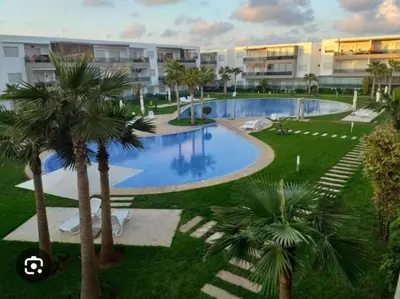 a swimming pool with palm trees in front of a building at Appartement Moderne 2 chambres à Blanca Beach Sidi Rahal in Sidi Rahal
