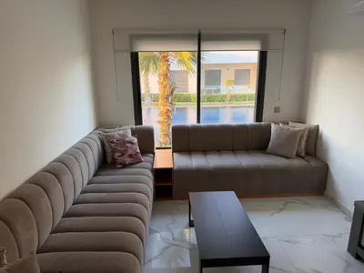 a living room with a couch and a table and a palm tree at Superbe appartement 120 mc, Plage à pieds, 3 piscines & calme garanti - Sidi Rahal Plage in Sidi Rahal