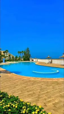 a large blue swimming pool with palm trees and a blue sky at Sea View Luxury Apartment Just Steps from the beach in Bouznika